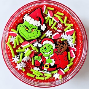 May include: A round container filled with red glittery slime topped with two Grinch-themed rubber charms, one showing a face in a Santa hat and the other a full-body figure with a sack, accented by white snowflake and lime green cylinder sprinkles.