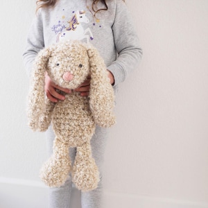 May include: A plush, beige bunny with long floppy ears and a pink nose is held by a person. The bunny has a textured, curly appearance. The person is wearing a gray sweatshirt with a unicorn graphic.