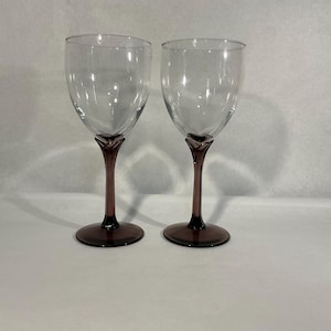 May include: Two clear glass wine glasses featuring deep purple stems and circular bases. The bowls are transparent and smooth, transitioning into a sculpted, petal-like design where the stem meets the base of the glass bowl.