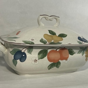 May include: A cream-colored ceramic casserole dish with a matching lid, featuring a speckled finish and a painted pattern of green leaves, white flowers, and various fruits including oranges, pears, and dark blue plums with gray trim.