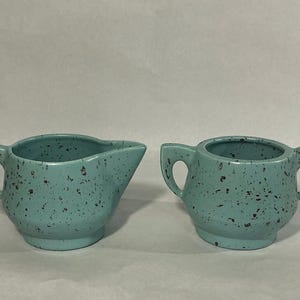 May include: A ceramic creamer pitcher and a matching sugar bowl, both featuring a light teal blue base color with a dark brown speckled pattern. The creamer has a single handle and a pouring spout, while the sugar bowl has two side handles.