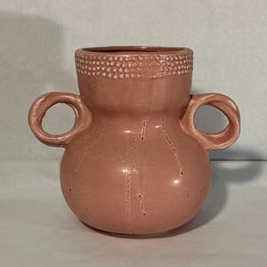 May include: A dusty rose ceramic vase with a rounded body and two side handles. The upper rim features a textured band of small raised dots, while the smooth surface of the vase is decorated with vertical drip-like glaze patterns.