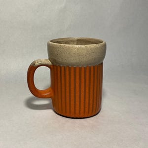 May include: A ceramic mug featuring a two-tone design with a speckled cream-colored upper section and a ribbed, vertically textured orange lower body. The mug includes a matching orange handle and a smooth, rounded rim, showcasing a distinct contrast.