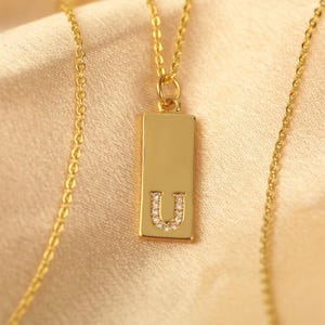 May include: Gold-colored necklace with a rectangular pendant. The pendant features the letter "U" encrusted with small, clear gemstones. The chain and pendant have a shiny finish, and the background is a soft, neutral color.