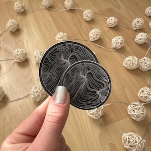 May include: Two round black stickers with white swirling line designs are held in front of a wooden surface. A string of white woven ball lights is in the background, creating a decorative effect.