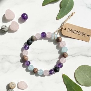 Könnte beinhalten: Ein handgefertigtes Armband mit Edelsteinperlen in Rosa, Lila, Braun und Blau. Das Armband wird mit losen Edelsteinen und einem Anhänger mit der Aufschrift "HANDMADE" präsentiert. Eukalyptusblätter sind um das Armband angeordnet.