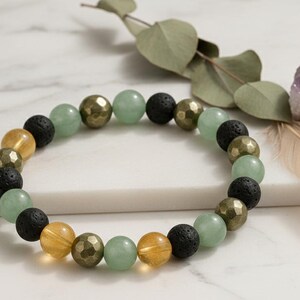 Könnte beinhalten: Ein Armband mit einer Mischung aus grünen, goldenen, schwarzen und gelben Perlen. Das Armband wird mit Eukalyptus, einem lila Kristall und einer Feder auf einer weißen Marmoroberfläche präsentiert.