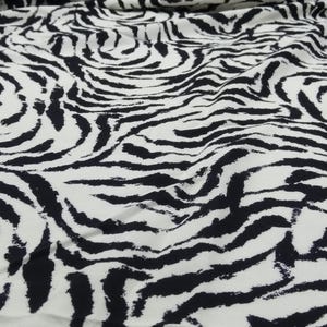 Pode incluir: Tecido branco com um padrão arrojado de listras de zebra pretas. O tecido está drapeado, mostrando sua textura e caimento. Ideal para vestuário, decoração de casa ou projetos de artesanato.