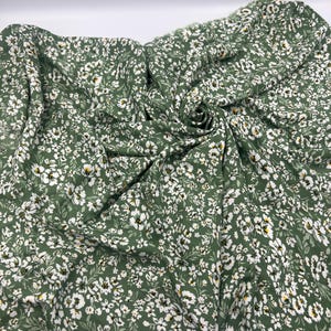 May include: Green fabric with a delicate floral pattern. The fabric is a deep green color, covered in small white flowers with yellow centers and small green leaves. The fabric is draped and folded, showing its texture and flow.