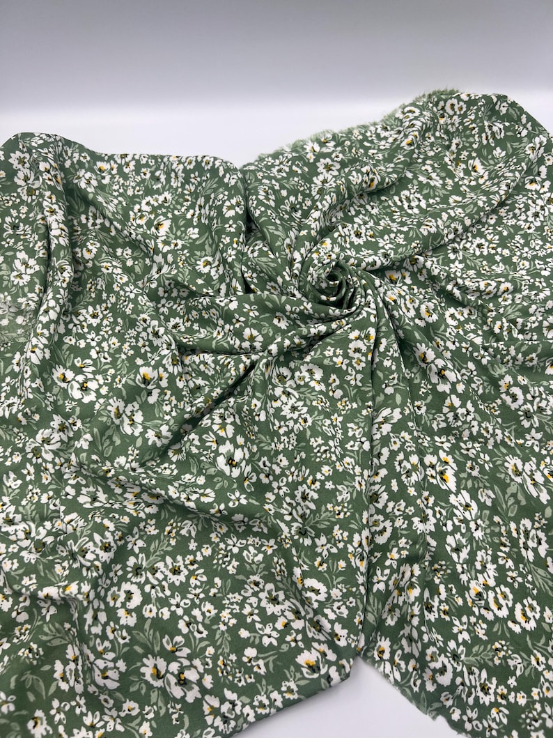May include: Green fabric with a delicate floral pattern. The fabric is a deep green color, covered in small white flowers with yellow centers and small green leaves. The fabric is draped and folded, showing its texture and flow.