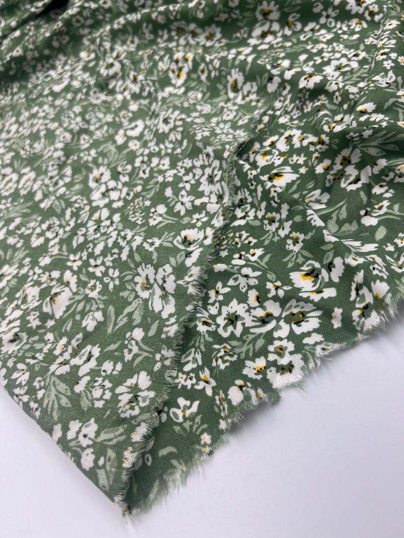 May include: Close-up of a green fabric with a floral pattern. The fabric is covered in small white flowers with yellow centers and green leaves. The edges of the fabric are frayed.