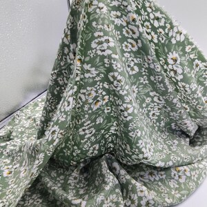 May include: A piece of green fabric with a delicate floral pattern. The fabric is covered in small white flowers with yellow centers, creating a fresh and natural look. The fabric is soft and flowing, suitable for clothing or home decor.