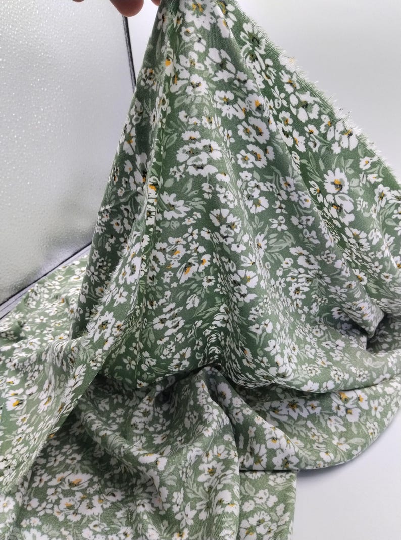 May include: A piece of green fabric with a delicate floral pattern. The fabric is covered in small white flowers with yellow centers, creating a fresh and natural look. The fabric is soft and flowing, suitable for clothing or home decor.
