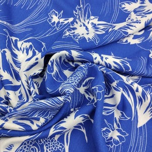 May include: A vibrant blue fabric featuring a repeating white floral pattern with intricate line work and curved details. The material is gathered in soft folds, showcasing the contrast between the bold blue background and the white botanical print.
