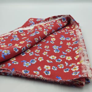 K&ouml;nnte beinhalten: Roter Blumenstoff mit einem sich wiederholenden Muster aus kleinen blauen und wei&szlig;en Blumen. Der Stoff hat einen ausgefransten Rand und ist auf sich selbst gefaltet. Der Stoff ist wahrscheinlich Baumwolle oder ein &auml;hnliches Material.
