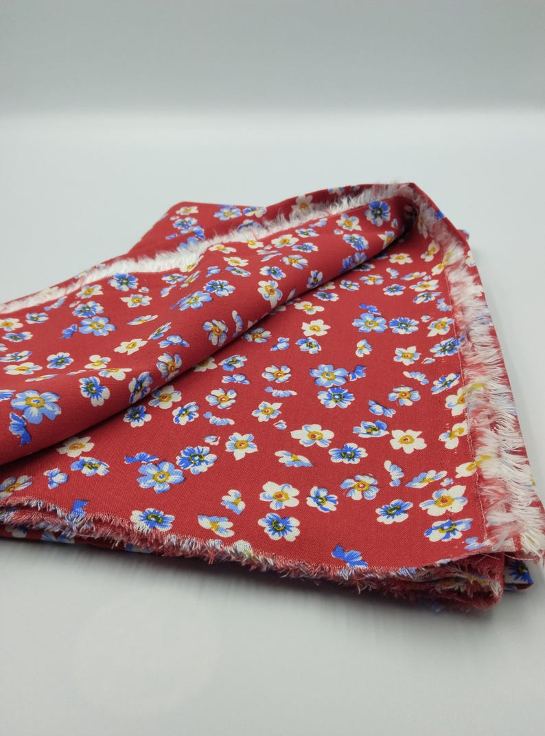 K&ouml;nnte beinhalten: Roter Blumenstoff mit einem sich wiederholenden Muster aus kleinen blauen und wei&szlig;en Blumen. Der Stoff hat einen ausgefransten Rand und ist auf sich selbst gefaltet. Der Stoff ist wahrscheinlich Baumwolle oder ein &auml;hnliches Material.