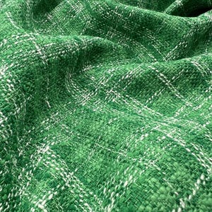 May include: Close-up of a textured green fabric with a woven pattern. White threads are interwoven throughout, creating a subtle grid-like design. The fabric appears soft and pliable, with visible folds and shadows.