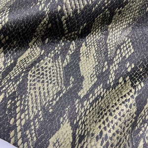Peut inclure: Un tissu présentant un motif répétitif de peau de serpent dans des tons gris anthracite foncé et vert olive sourd. Le matériau affiche une surface texturée avec un imprimé d'écailles détaillé qui crée une profondeur visuelle sur tout le textile.