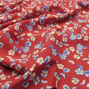 K&ouml;nnte beinhalten: Nahaufnahme eines roten Stoffes mit einem sich wiederholenden Blumenmuster. Die kleinen Blumen haben wei&szlig;e Bl&uuml;tenbl&auml;tter, gelbe Zentren und blaue Akzente. Der Stoff ist drapiert und zeigt Falten und Schatten, was einen strukturierten Eindruck erzeugt.