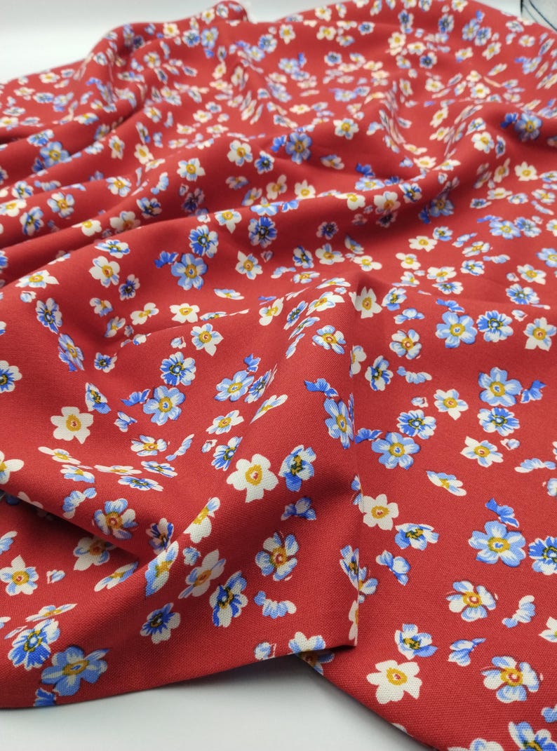 K&ouml;nnte beinhalten: Nahaufnahme eines roten Stoffes mit einem sich wiederholenden Blumenmuster. Die kleinen Blumen haben wei&szlig;e Bl&uuml;tenbl&auml;tter, gelbe Zentren und blaue Akzente. Der Stoff ist drapiert und zeigt Falten und Schatten, was einen strukturierten Eindruck erzeugt.