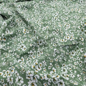 May include: Close-up of a green fabric with a delicate floral pattern. The fabric is covered in small white flowers with yellow centers, creating a soft and inviting texture. The fabric is likely used for clothing or home decor.
