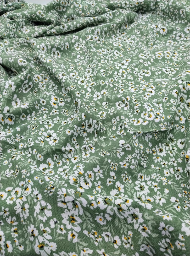 May include: Close-up of a green fabric with a delicate floral pattern. The fabric is covered in small white flowers with yellow centers, creating a soft and inviting texture. The fabric is likely used for clothing or home decor.