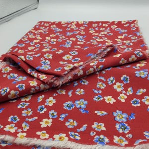 K&ouml;nnte beinhalten: Roter Stoff mit einem sich wiederholenden Blumenmuster aus kleinen wei&szlig;en und blauen Bl&uuml;ten. Der Stoff ist gefaltet und zeigt die ausgefransten Kanten des Materials. Die Bl&uuml;ten haben gelbe Zentren.