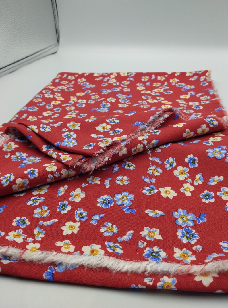 K&ouml;nnte beinhalten: Roter Stoff mit einem sich wiederholenden Blumenmuster aus kleinen wei&szlig;en und blauen Bl&uuml;ten. Der Stoff ist gefaltet und zeigt die ausgefransten Kanten des Materials. Die Bl&uuml;ten haben gelbe Zentren.