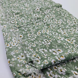 May include: Green fabric with a white floral pattern and frayed edges. The fabric is covered in small white flowers with yellow centers and green leaves. The fabric is folded over, showing the pattern and texture.