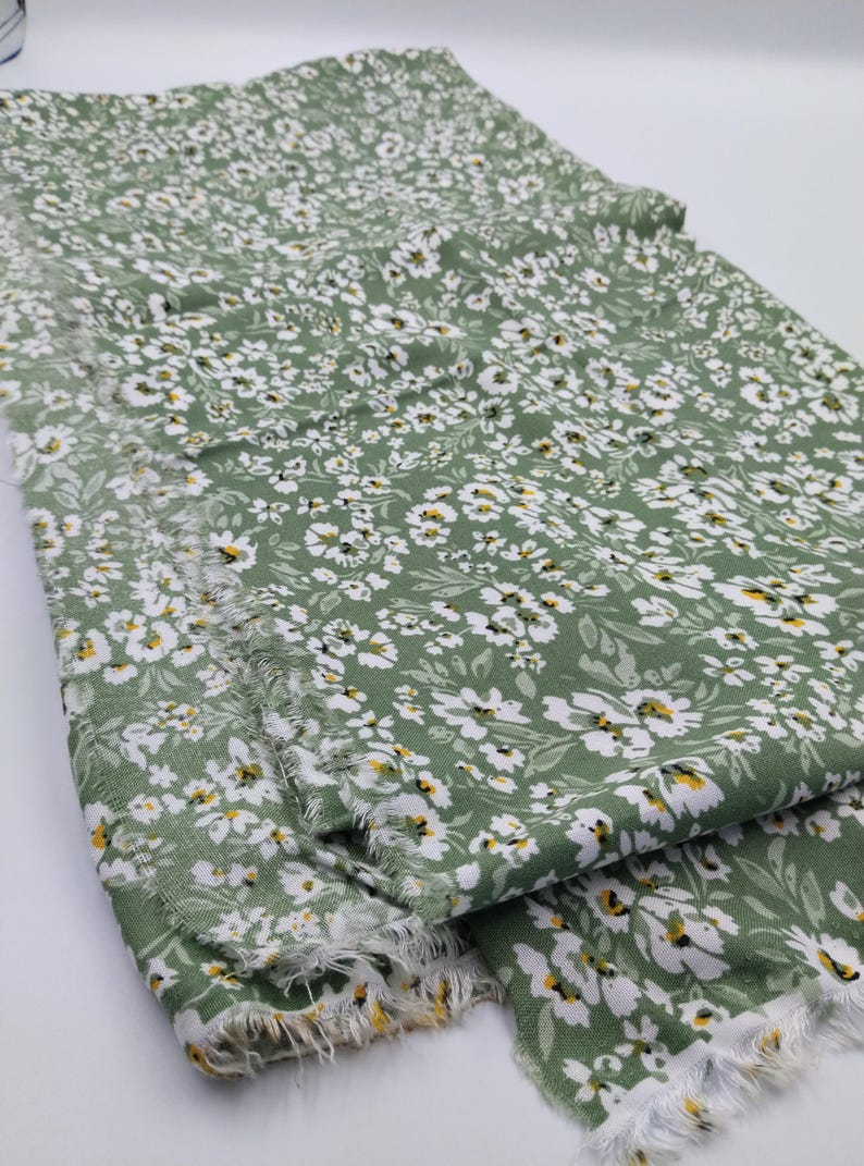 May include: Green fabric with a white floral pattern and frayed edges. The fabric is covered in small white flowers with yellow centers and green leaves. The fabric is folded over, showing the pattern and texture.