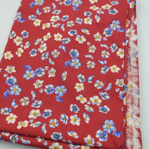 K&ouml;nnte beinhalten: Gefalteter roter Stoff mit einem sich wiederholenden Blumenmuster aus blauen, wei&szlig;en und gelben Bl&uuml;ten. Der Stoffrand hat einen Fransenbesatz. Geeignet f&uuml;r N&auml;h- oder Bastelprojekte.