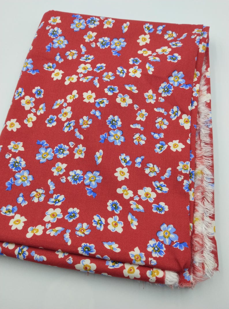 K&ouml;nnte beinhalten: Gefalteter roter Stoff mit einem sich wiederholenden Blumenmuster aus blauen, wei&szlig;en und gelben Bl&uuml;ten. Der Stoffrand hat einen Fransenbesatz. Geeignet f&uuml;r N&auml;h- oder Bastelprojekte.