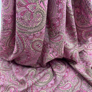 May include: Pink paisley fabric with a detailed pattern in shades of pink, beige, and brown. The fabric is draped, showing the texture and flow of the material. The design features a classic paisley motif.