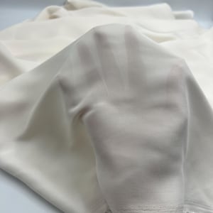 Può includere: Un pezzo di tessuto bianco sporco, semi-trasparente, con una consistenza liscia e opaca. Il materiale è drappeggiato in morbide pieghe, rivelando una qualità traslucida che permette di vedere la sagoma di una mano attraverso il tessuto.