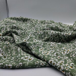 May include: Close-up of a green fabric with a delicate floral pattern. The fabric is covered in small white flowers with yellow centers, creating a soft, textured appearance. The fabric is draped, showing its folds and flow.