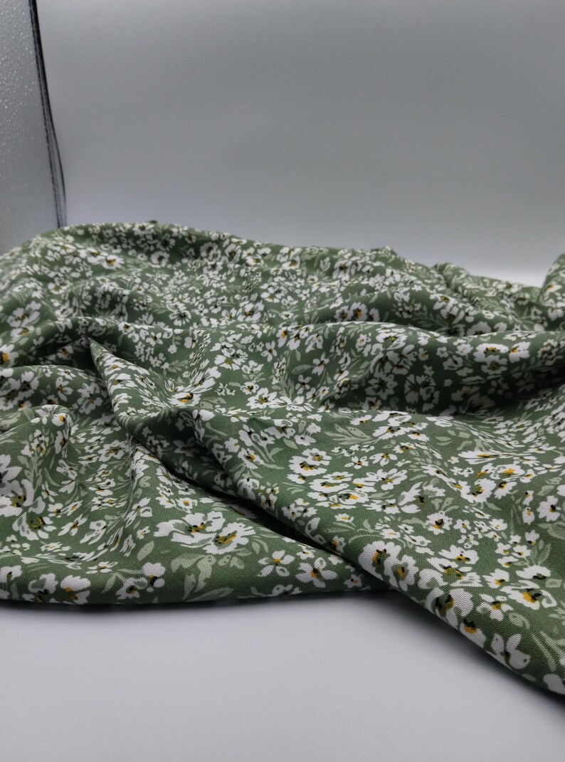 May include: Close-up of a green fabric with a delicate floral pattern. The fabric is covered in small white flowers with yellow centers, creating a soft, textured appearance. The fabric is draped, showing its folds and flow.