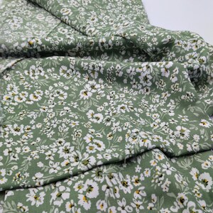 May include: Green fabric with a white floral pattern. The fabric is covered in small white flowers with yellow centers and green leaves. The fabric is folded over, showing the pattern and the frayed edge.