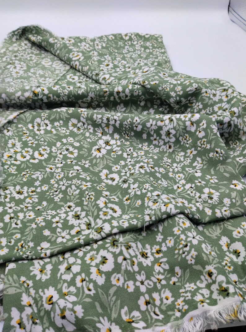 May include: Green fabric with a white floral pattern. The fabric is covered in small white flowers with yellow centers and green leaves. The fabric is folded over, showing the pattern and the frayed edge.