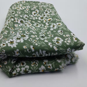 May include: Folded fabric with a green background and a white floral pattern. The fabric is a light green color with small white flowers and yellow centers. The fabric is folded over on itself.