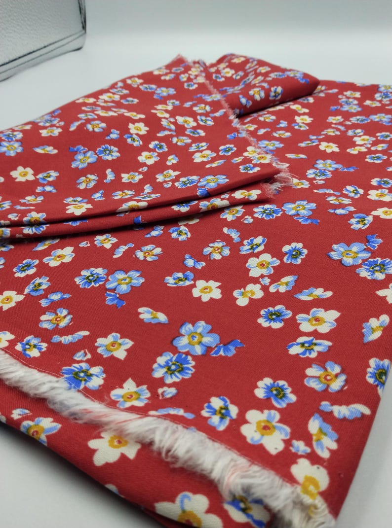K&ouml;nnte beinhalten: Roter Stoff mit floralem Muster. Der Stoff ist mit kleinen blauen, wei&szlig;en und gelben Blumen bedeckt. Die Kanten des Stoffes sind ausgefranst und zeigen die rohe Kante des Materials. Der Stoff ist gefaltet.