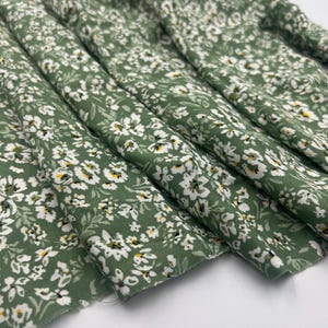 May include: Close-up of a folded fabric with a floral pattern. The fabric is a muted green color, covered with small white flowers and tiny yellow accents. The fabric is likely cotton or a similar material, suitable for sewing projects.