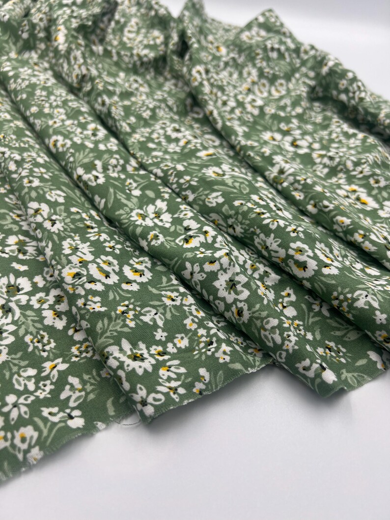 May include: Close-up of a folded fabric with a floral pattern. The fabric is a muted green color, covered with small white flowers and tiny yellow accents. The fabric is likely cotton or a similar material, suitable for sewing projects.