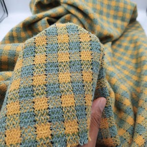 Op de afbeelding: Een geweven stof met een ruitpatroon in tinten mosterdgeel en saliegroen. Het textiel heeft een zichtbare rasterstructuur met dunne, donkere verticale lijnen die door het weefsel lopen, wat zorgt voor een getextureerd uiterlijk.