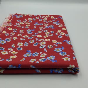 K&ouml;nnte beinhalten: Gefalteter roter Stoff mit Blumenmuster. Der Stoff ist mit kleinen blauen und wei&szlig;en Blumen bedeckt. Der Rand des Stoffes hat eine Fransenborte. Der Stoff ist auf sich selbst gefaltet.