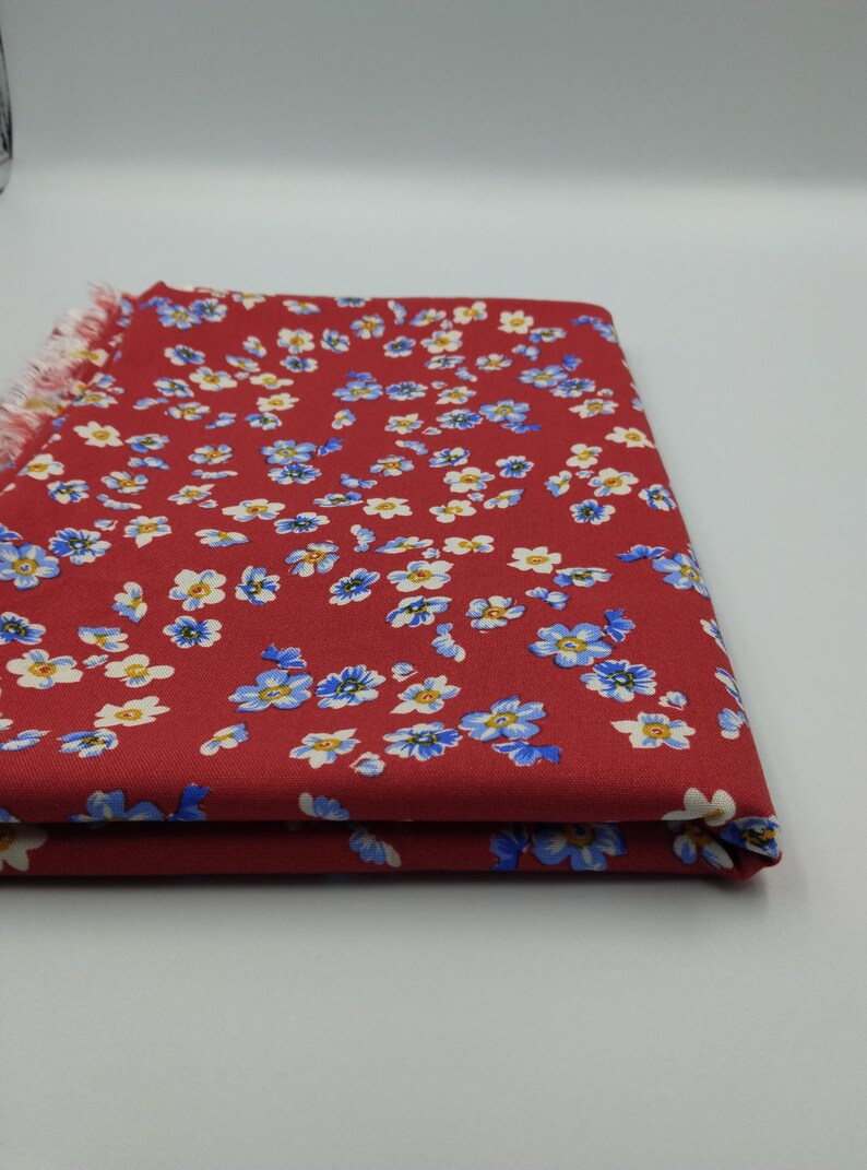 K&ouml;nnte beinhalten: Gefalteter roter Stoff mit Blumenmuster. Der Stoff ist mit kleinen blauen und wei&szlig;en Blumen bedeckt. Der Rand des Stoffes hat eine Fransenborte. Der Stoff ist auf sich selbst gefaltet.