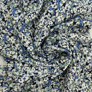 May include: Close-up of a fabric with a floral pattern. The fabric features small white, blue, and green flowers against a dark navy blue background. The fabric is gathered and swirled, showing the texture and drape of the material.