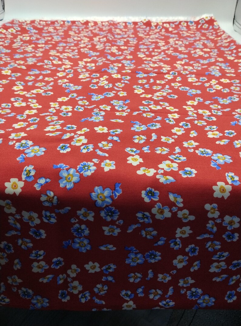 K&ouml;nnte beinhalten: Roter Stoff mit einem Muster aus kleinen blauen, wei&szlig;en und gelben Blumen. Das florale Design ist gleichm&auml;&szlig;ig &uuml;ber die Stoffoberfl&auml;che verteilt und erzeugt ein dichtes und farbenfrohes Muster.