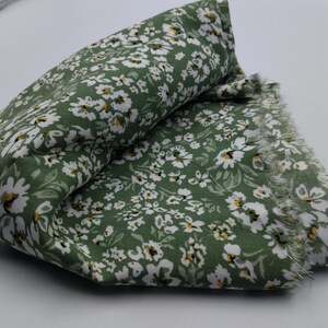 May include: Folded green fabric with a white floral pattern and frayed edges. The fabric has a dense pattern of small white flowers with yellow centers, set against a solid green background. The fabric is folded over on itself.