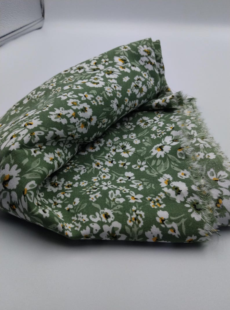 May include: Folded green fabric with a white floral pattern and frayed edges. The fabric has a dense pattern of small white flowers with yellow centers, set against a solid green background. The fabric is folded over on itself.