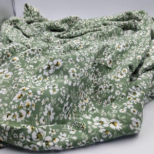 May include: A folded piece of fabric with a repeating floral pattern. The fabric is a muted green color, covered in small white flowers with yellow centers. The fabric appears soft and lightweight, suitable for clothing or home decor.